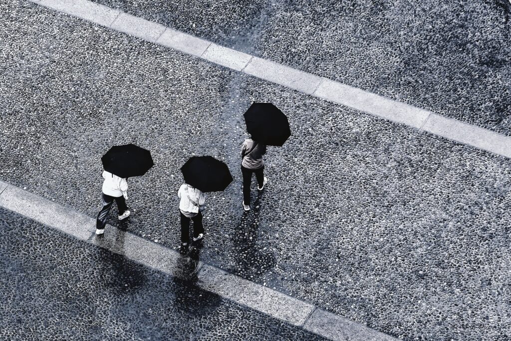 Drei Personen mit dunklen Regenschirmen auf nassem Straßenbelag, von oben fotografiert.