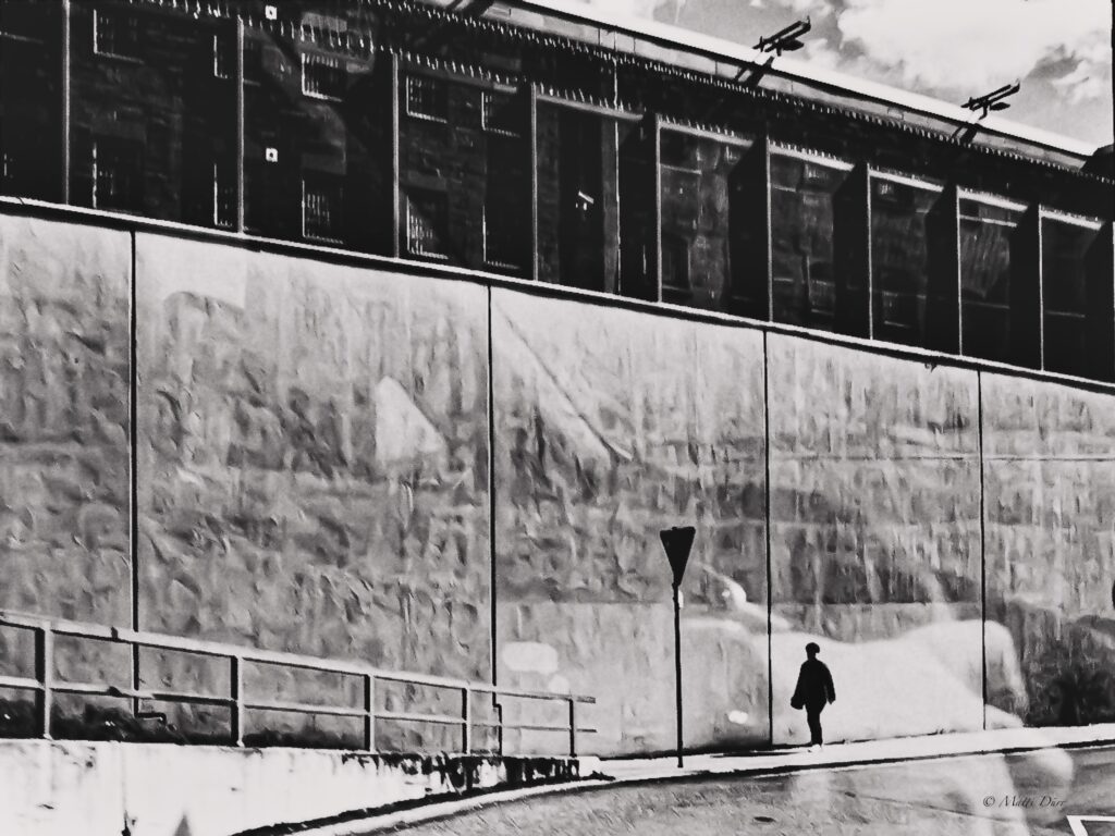 Fotografía en blanco y negro de una persona sola en una acera, junto a un muro alto y un edificio alargado.